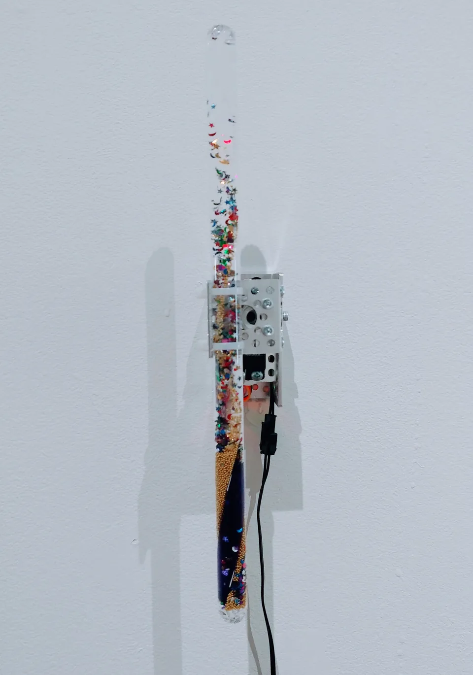 A long transparent wand filled with metallic stars, crescent shapes, and gold-colored beads is attached to a motor near its middle.