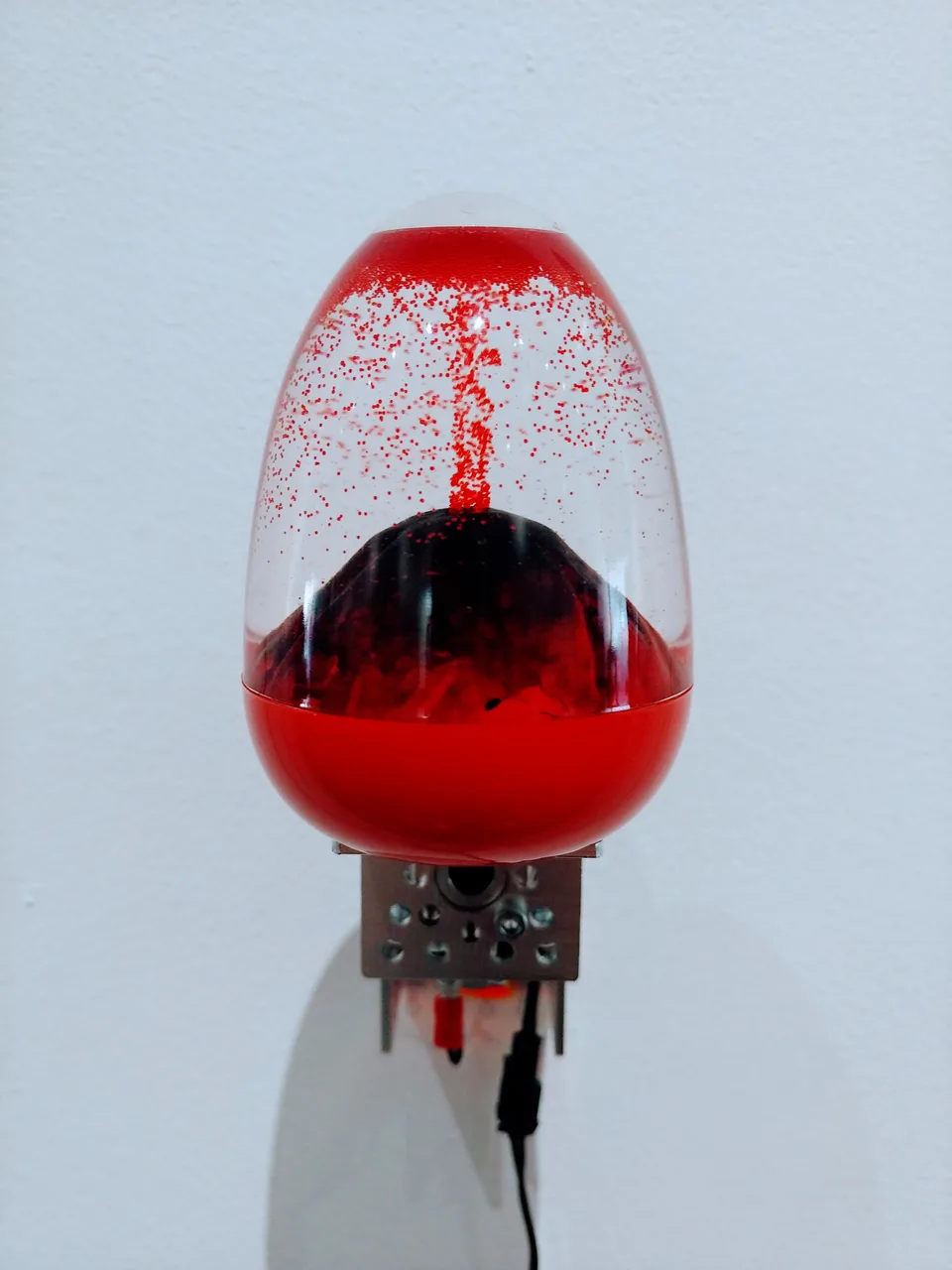 A transparent egg-shaped toy with a red base contains a miniature volcano and red particles above a small motorized mechanism.