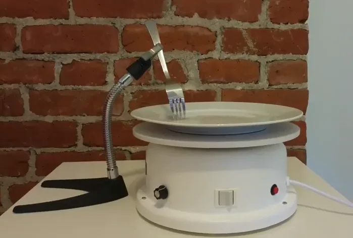 A flexible metal arm holds a fork against a plate on a motorized base