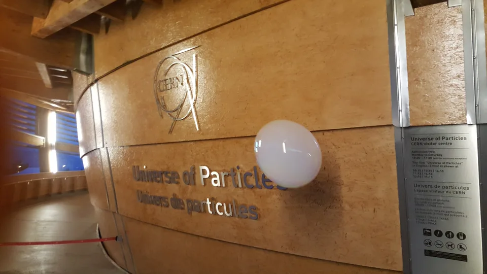 A large pale bubble floats in front of the CERN emblem and Universe of Particles signage on a curved wooden wall.
