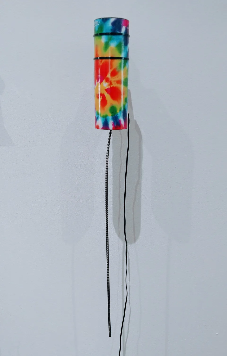 A tube patterned in red, yellow, blue, and green has a long metal spring hanging beneath it and wiring at its side.