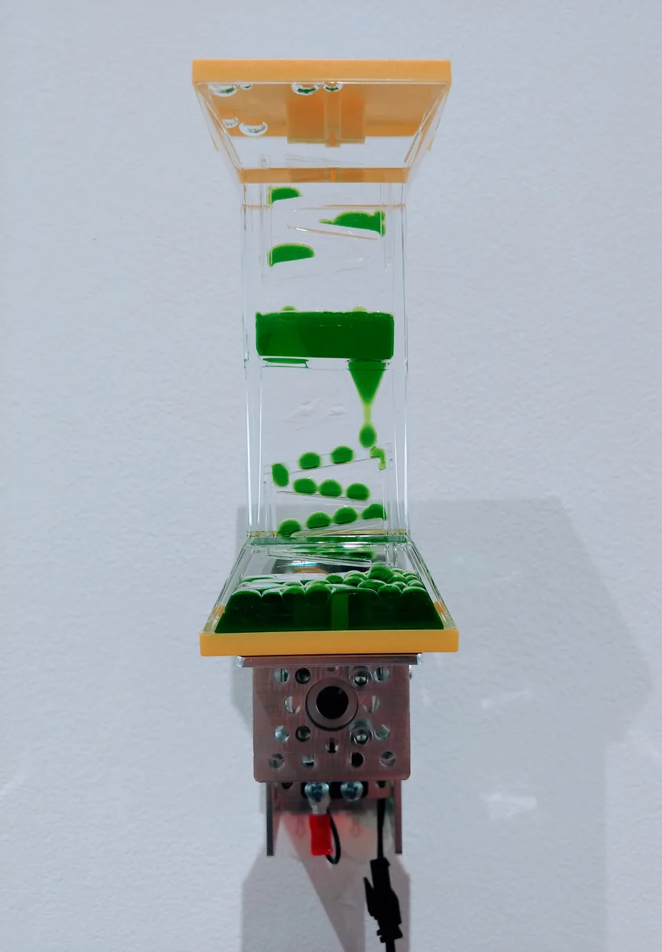 A rectangular transparent toy with yellow end caps and descending green liquid droplets is mounted above a motor.