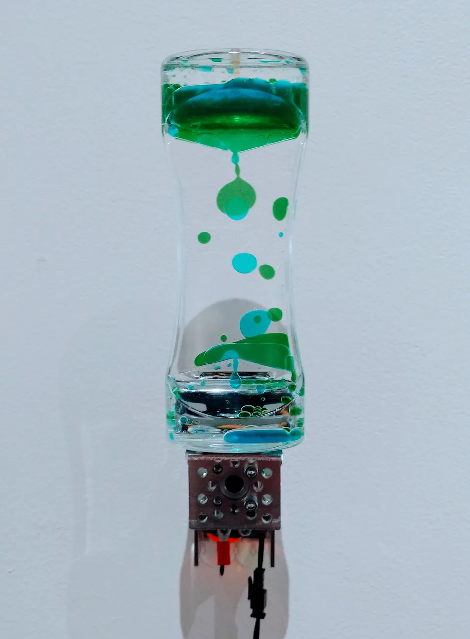 A curved transparent hourglass-shaped toy containing green liquid droplets is attached to a motor.