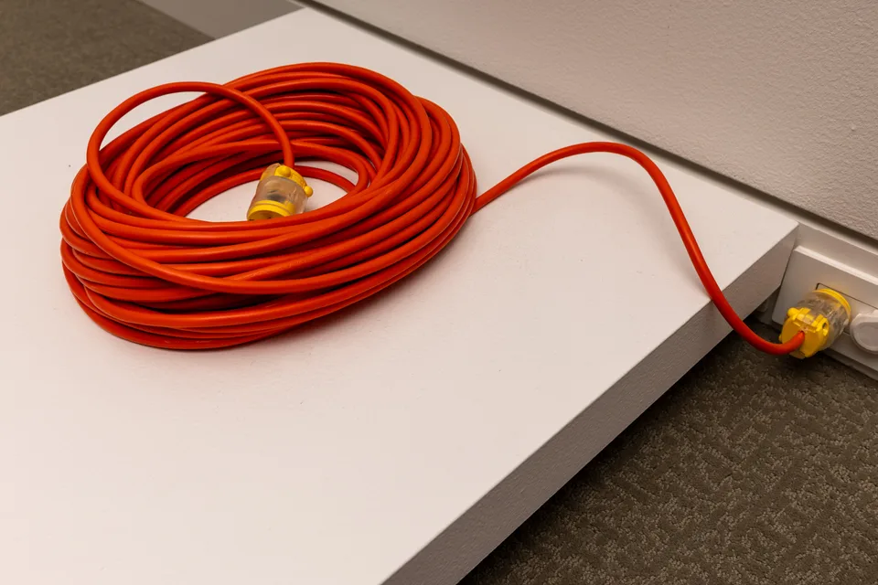 An orange extension cord is coiled on a low white plinth, with one plug extending toward the wall.