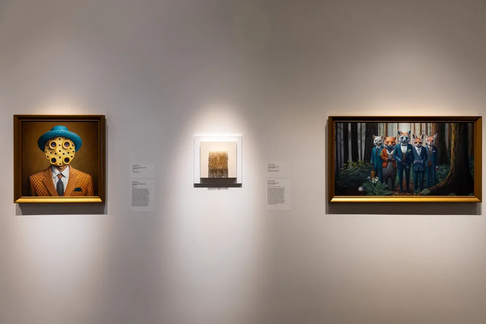 Three works on a white gallery wall: a yellow-faced portrait, a small pin-covered canvas, and a painting of fox-headed figures.